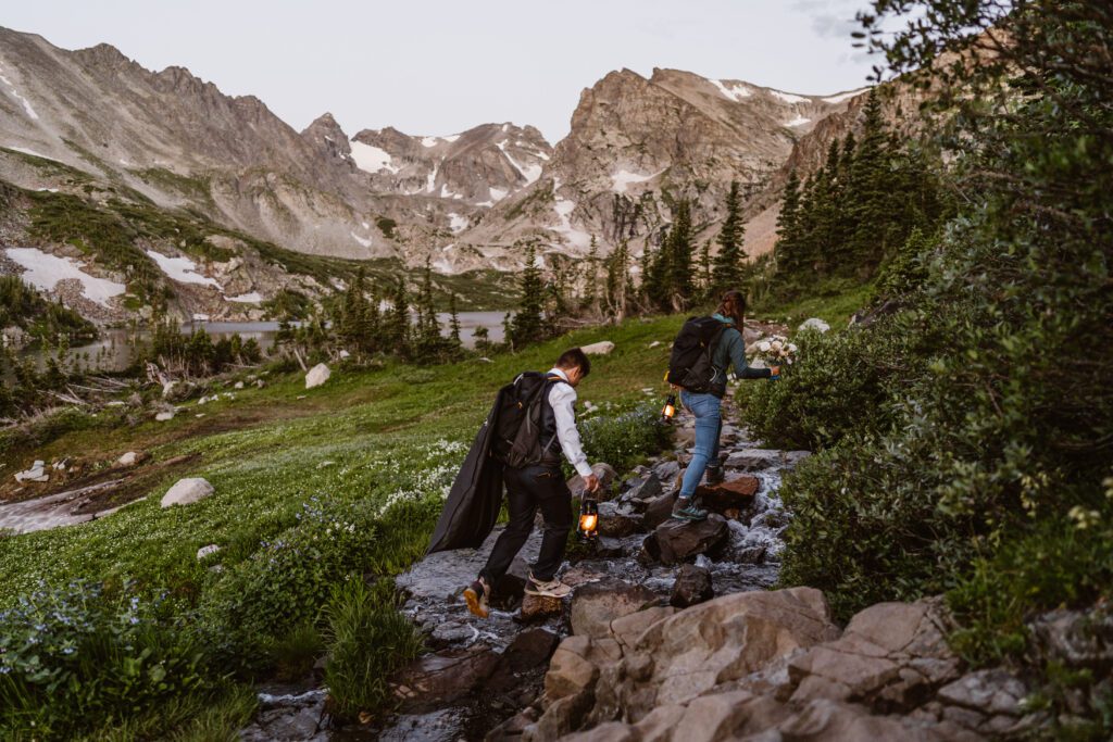 Wedding couple hikes over a stream crossing at sunrise for their Lake Isabelle elopement 