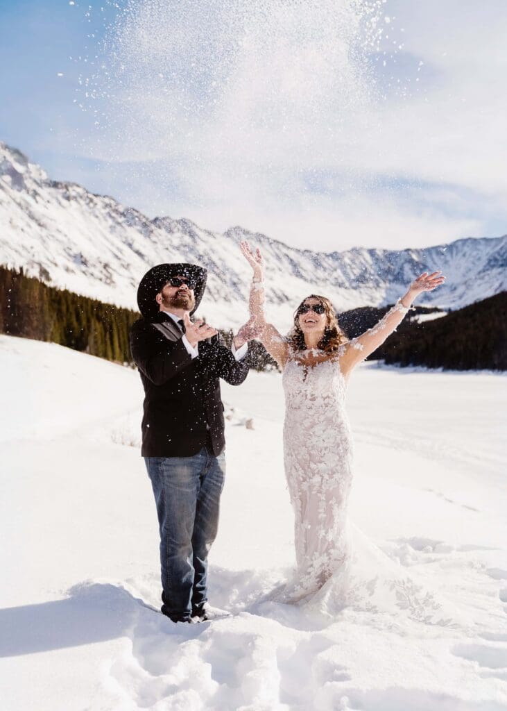 A bride and groom wearing sunglasses and laughing while throwing snow in the air during their adventurous winter elopement.