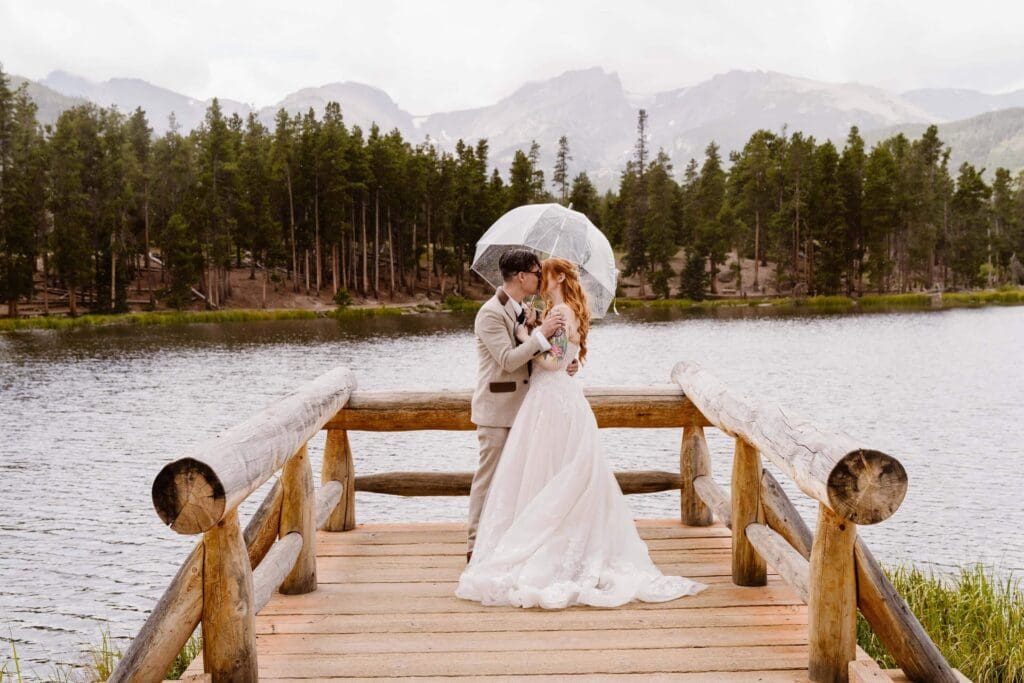 A bride and groom sharing a quiet moment under a clear umbrella on a wooden dock at Sprague Lake in Rocky Mountain National Park.