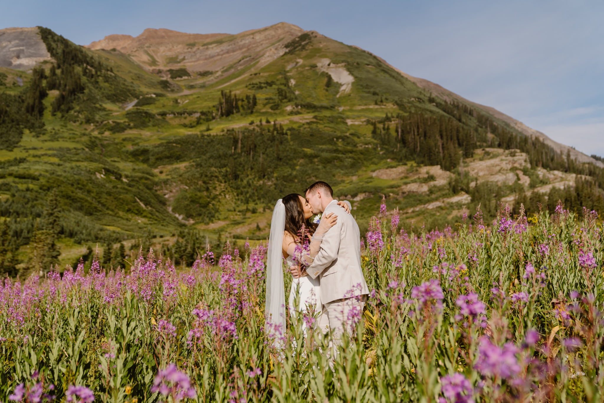 A bride and groom share a kiss in a vibrant field of wildflowers during their Crested Butte, Colorado elopement. Surrounded by tall purple blooms and dramatic green mountains under a sunny blue sky, the couple stands wrapped in an intimate embrace, radiating love and joy.
