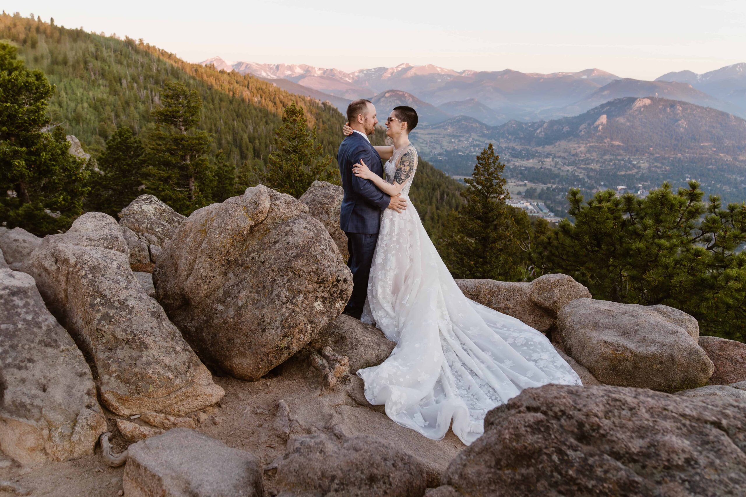 A bride and groom kiss on a rocky overlook with the Rocky Mountain peaks glowing at sunset in Estes Park, Colorado.
