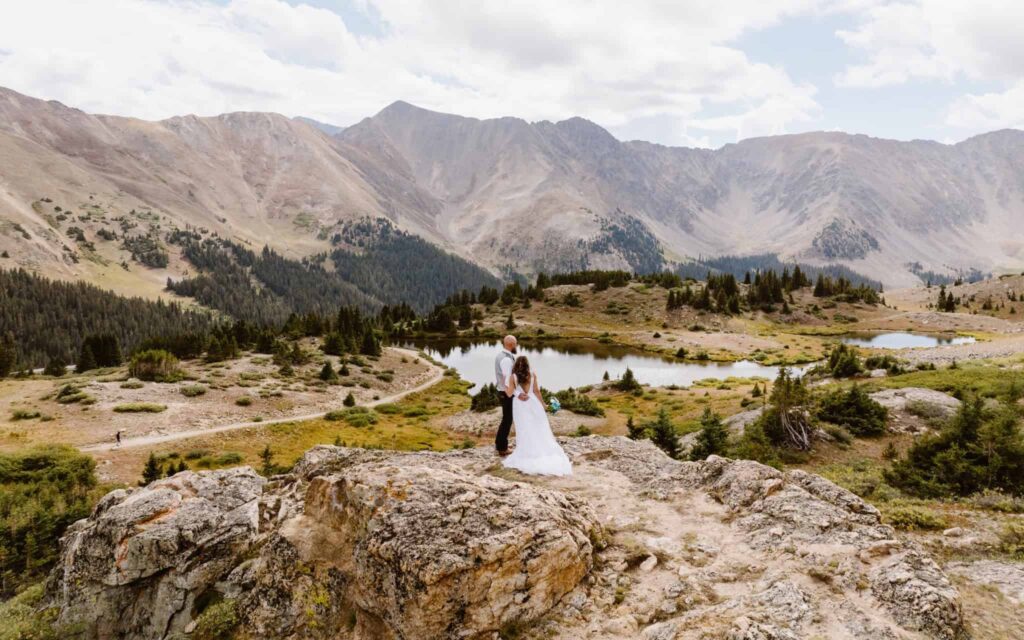 Where to elope in Breckenridge: Bride and groom stand on a rocky outcrop at Loveland Pass, Colorado, overlooking alpine lakes, evergreen forests, and towering mountain peaks.