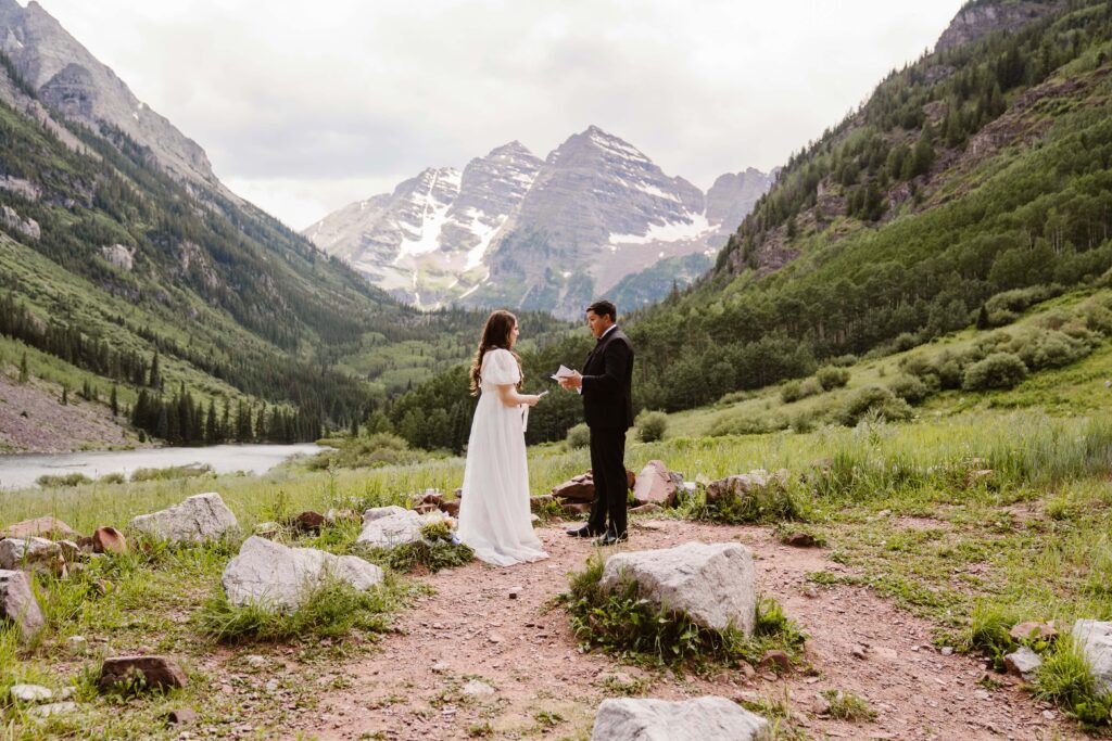 Bride and groom exchange vows during an intimate elopement ceremony at Maroon Bells Amphitheater, surrounded by lush greenery, towering pine trees, and dramatic snow-capped Maroon Bells peaks in the background.