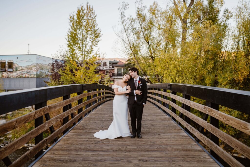 A bride and groom embrace on a rustic wooden bridge in Steamboat Springs, Colorado, surrounded by golden autumn trees. The bride rests her head on the groom’s shoulder in a fitted off-the-shoulder gown, while the groom wears a classic black suit with a floral tie and boutonniere. A colorful mural and cozy town buildings peek out in the background.