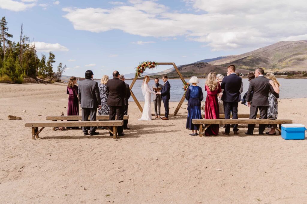 Where to elope in Breckenridge: Wedding ceremony at Windy Point Campground in Colorado, with the couple standing under a wooden hexagon arch adorned with flowers on a sandy lakeshore surrounded by mountains.