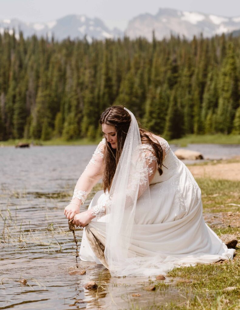 Bride kneeling by the water rinsing her wedding dress after hiking at their Brainard Lake elopement