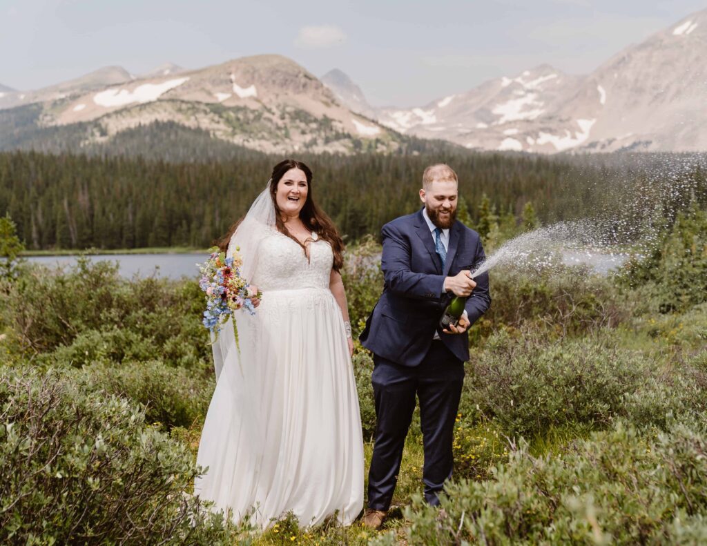 Groom popping a bottle of champagne while bride laughs holding her bouquet during their Brainard Lake elopement