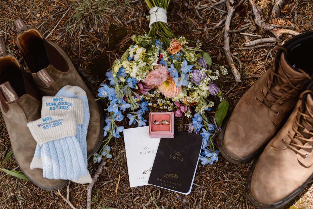 Wedding flatlay with wildflower bouquet, vow books, rings, boots, and custom socks at Brainard Lake elopement