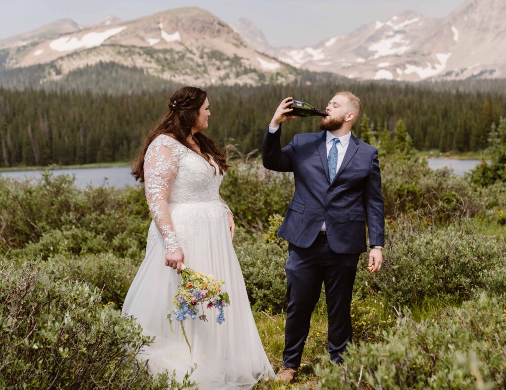 Groom drinking from a champagne bottle while bride watches at their Brainard Lake elopement