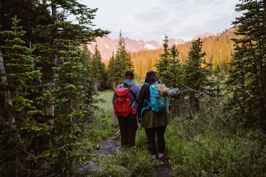 Couple hiking through alpine forest at sunrise with backpacks on their way to their Lake Isabelle elopement
