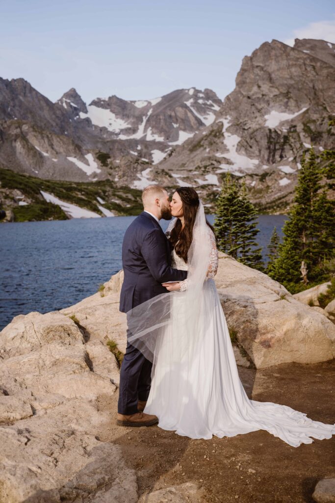 Bride and groom sharing a romantic kiss on the rocky lakeshore surrounded by alpine peaks at their Lake Isabelle elopement