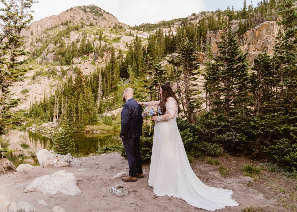 Bride tapping groom on the shoulder during their first look at the edge of Lake Isabelle surrounded by mountains