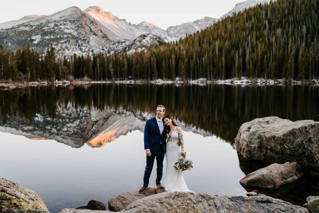 Bride and groom standing on lakeside rocks at sunset, Bear Lake perfectly reflecting the surrounding pine forest and alpenglow peaks with Colorado Elopement Photographer and Videographer Team Alpine Vows
