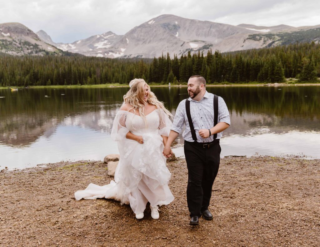 Playful couple walking hand in hand along the shore of Brainard Lake with reflections of rugged peaks and summer greenery behind them.