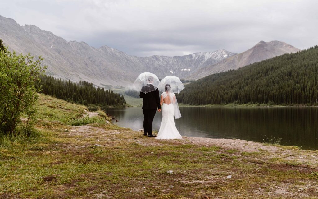 Couple holding clear umbrellas during a rainy-day elopement by a peaceful alpine lake surrounded by mountain peaks near Breckenridge with Breckenridge elopement photographer and videographer Alpine Vows.