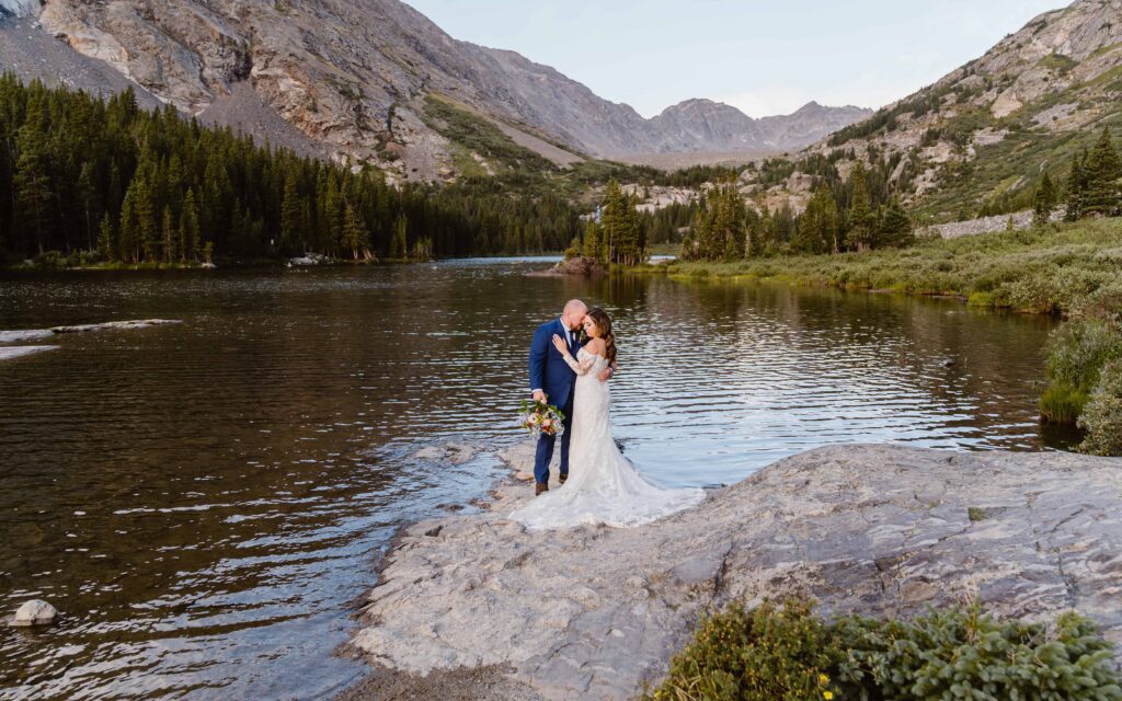 Bride and groom share a kiss on a rock beside an alpine lake with dramatic mountain peaks near Breckenridge.
