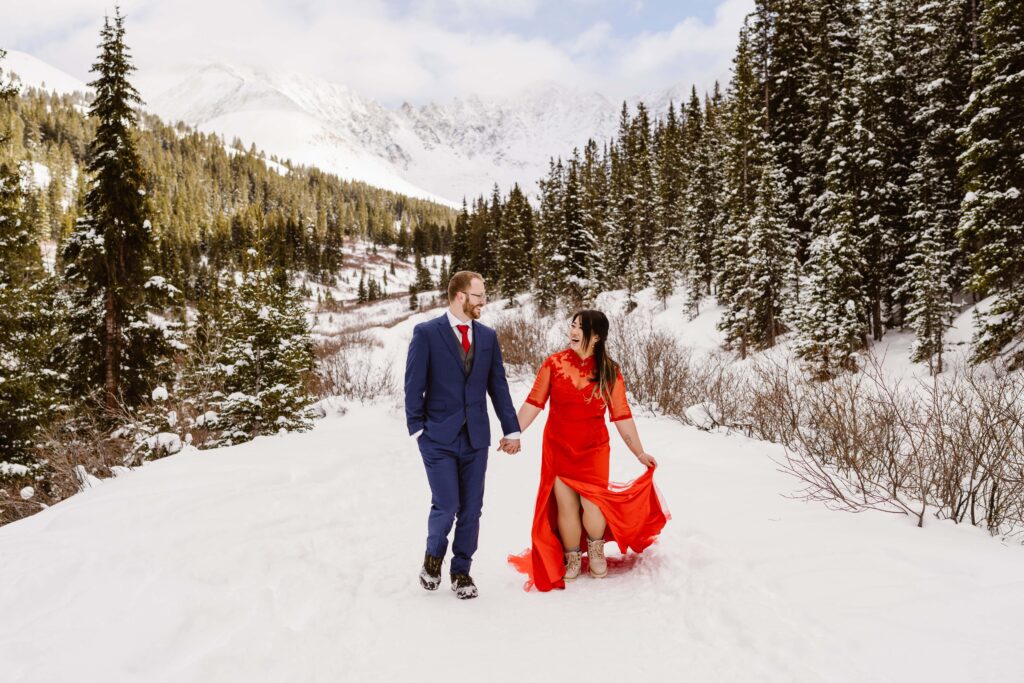 Bride in a bold red dress and groom in a blue suit walking hand in hand through snowy pines at Breckenridge, Colorado with Colorado Elopement Photographer and Videographer Team Alpine Vows