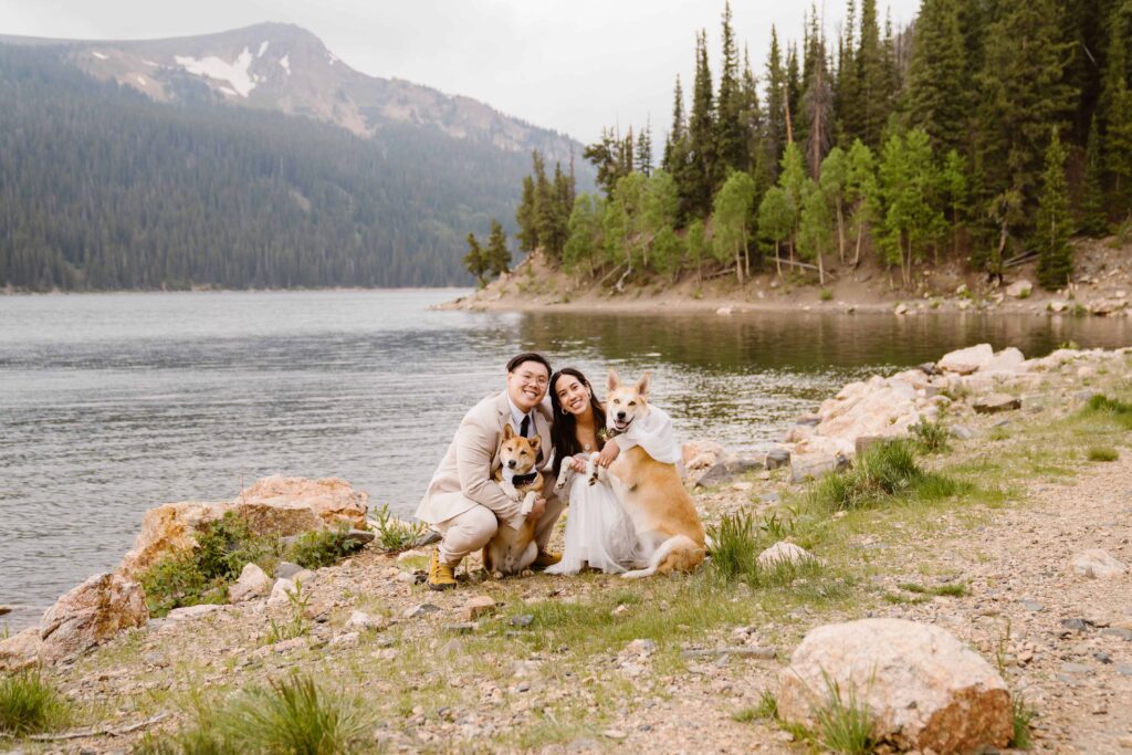 Bride and groom kneeling and smiling with their two dogs by a serene Colorado mountain lake.