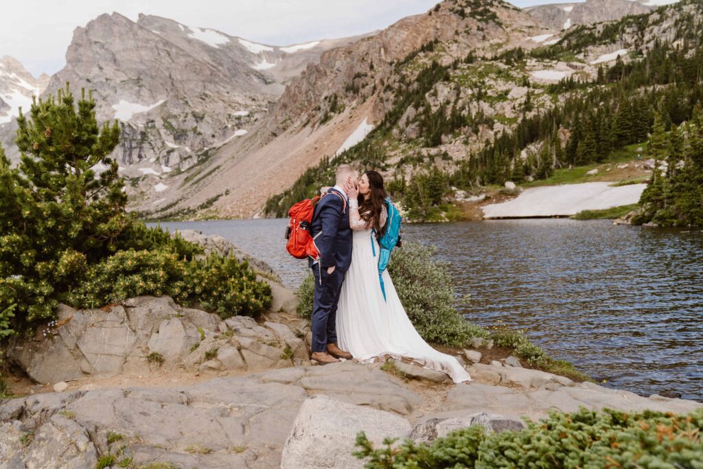 Adventure elopement couple kissing by Lake Isabelle with backpacks on, dramatic alpine cliffs and wild Colorado scenery behind them.