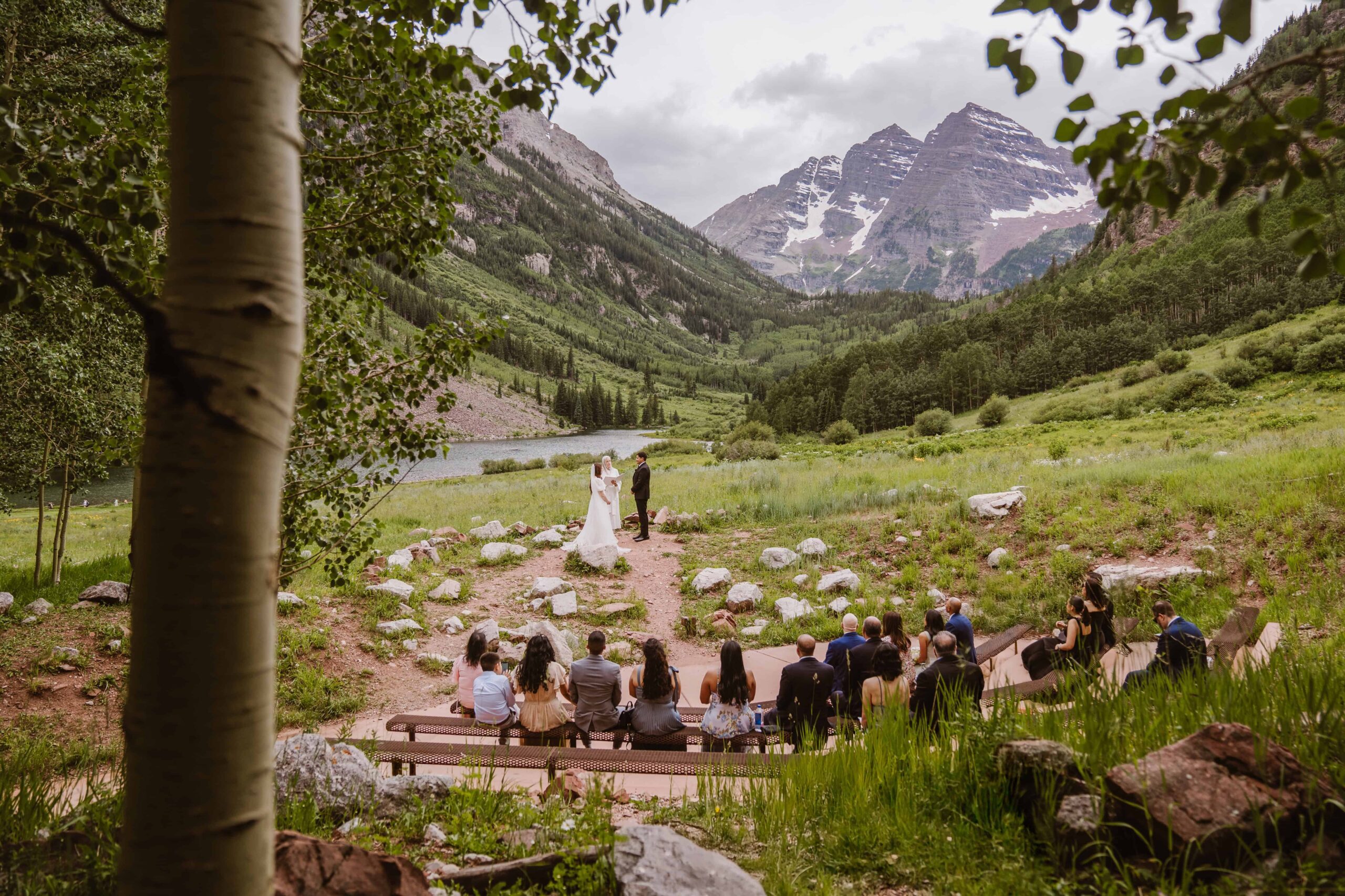 Wide view of a wedding ceremony at Maroon Bells Amphitheater with guests seated and mountain peaks rising in the background.