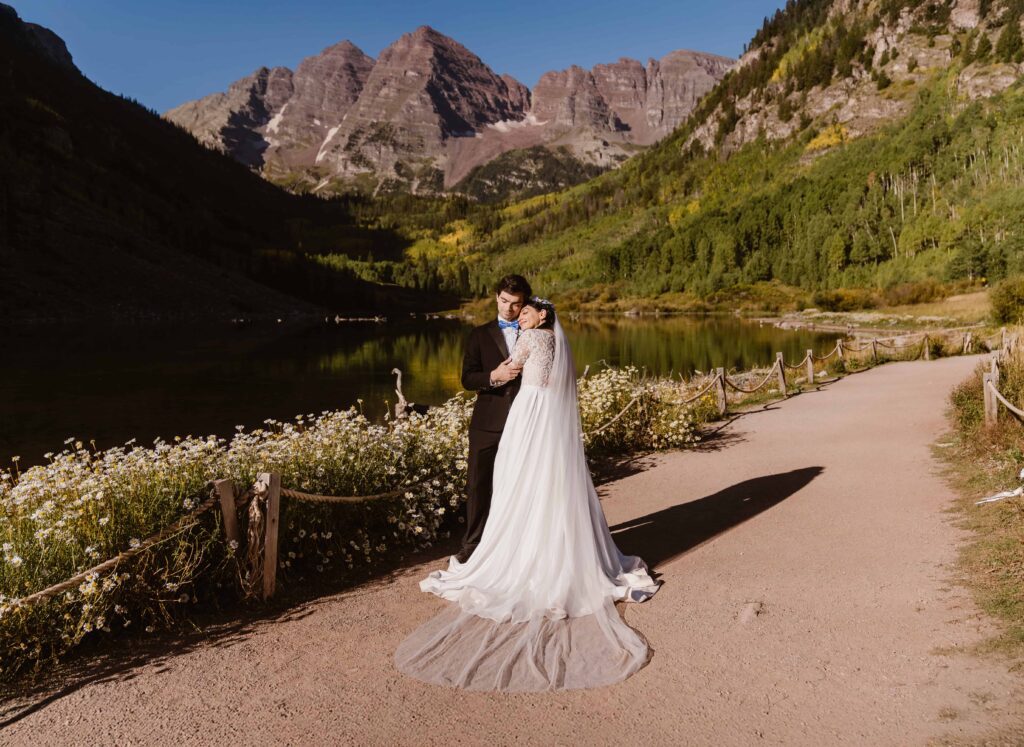 Couple embracing on a wildflower-lined trail at Maroon Bells Amphitheater with the iconic twin peaks and lake in the background with Maroon Bells Amphitheater elopement photographer Alpine Vows.