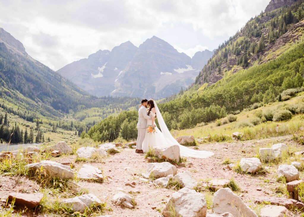 Bride and groom sharing a kiss on a rocky hillside at Maroon Bells with sweeping alpine peaks and lush green valley views.