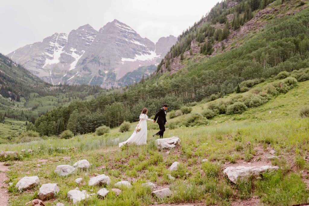 Newlyweds walking hand in hand across a lush green meadow with Maroon Bells peaks towering in the distance.