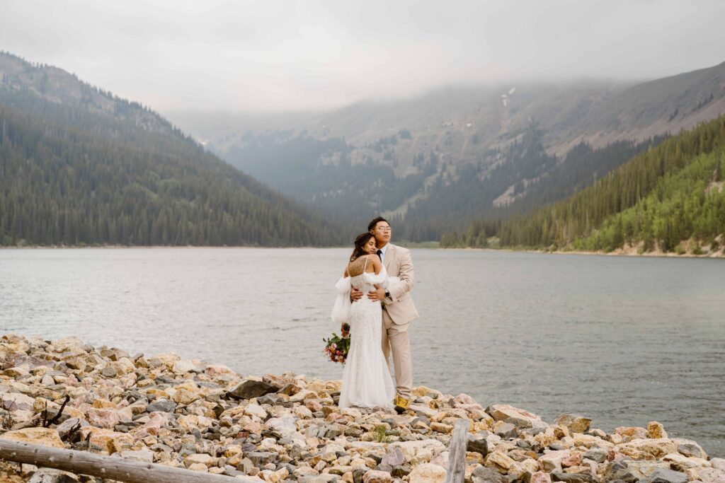 Couple holding each other on rocky shore with dramatic Colorado mountain lake views at their Airbnb wedding.