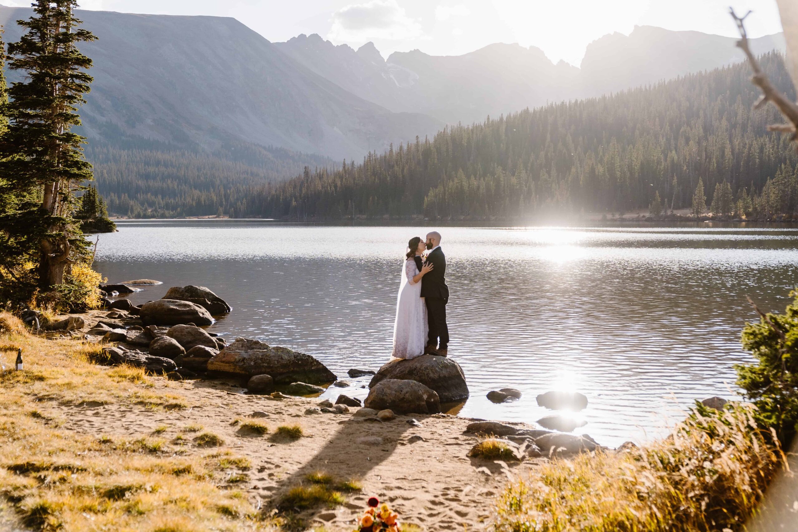How Much Does an Elopement Photographer Cost couple stands on a rock embracing each other with the lake and the mountains in the background and the sunset shining on the lake.