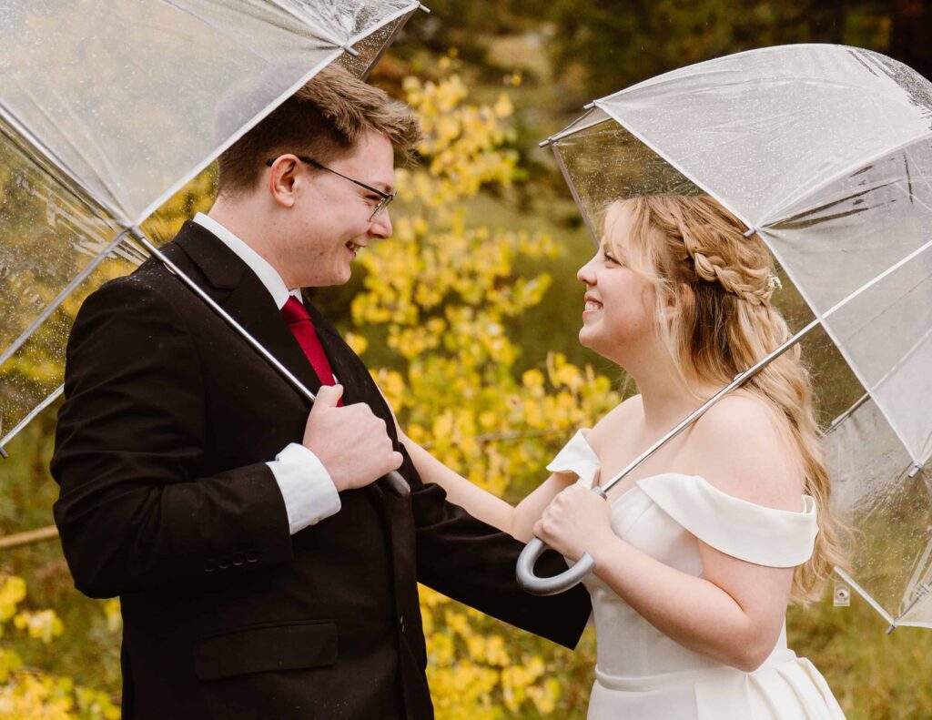 A close up of the couple sharing a joyful moment under umbrellas during their Estes Park elopement in peak fall color. Colorado Wedding and Portrait Photographer.