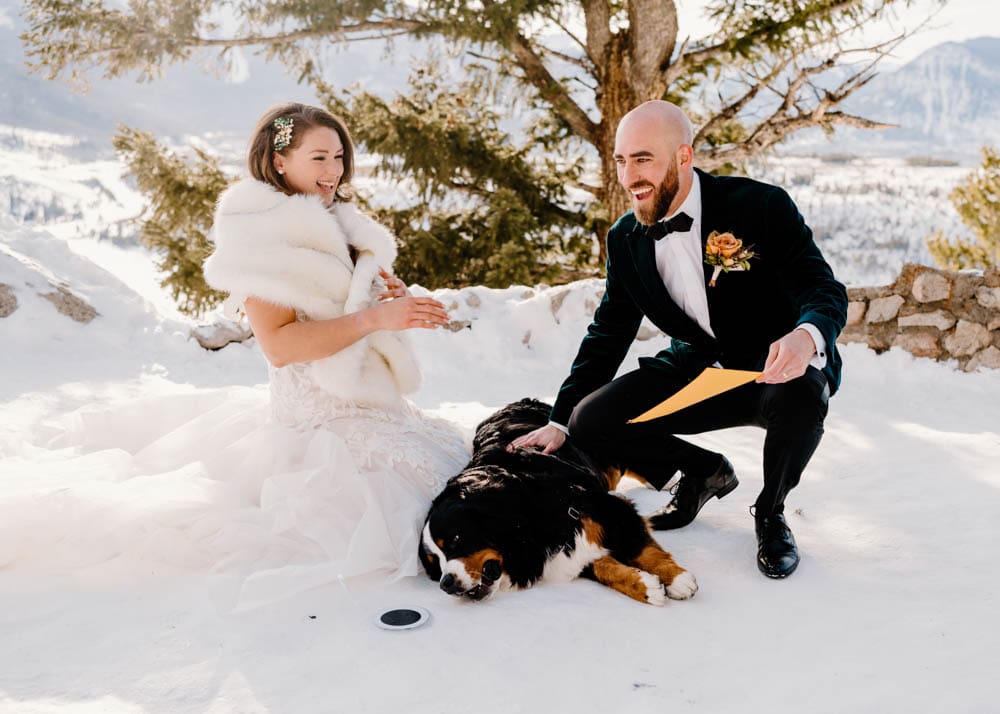 A couple is laughing in the snow as their Bernese Mountain Dog is about to elope at Sapphire Point Overlook by signing their marriage license with a paw print.