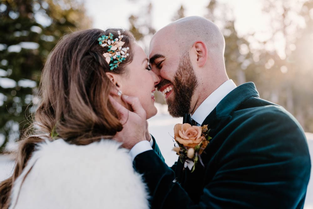 A close-up photograph of a bride and groom kissing during their winter micro-wedding, highlighting the bride's intricate teal floral hairpiece and the groom's matching boutonnière.