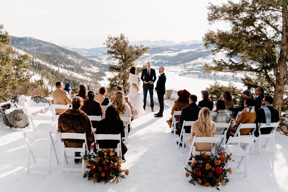 An overview shot of a winter micro-wedding ceremony setup at Sapphire Point with white chairs, orange and yellow floral arrangements, and panoramic mountain views.