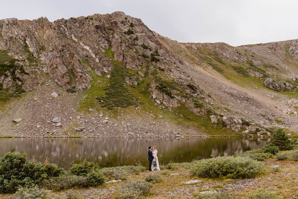 Couple stands together at the edge of an alpine lake during their elopement in Aspen, Colorado with Colorado elopement photographer and videographer alpine vows.